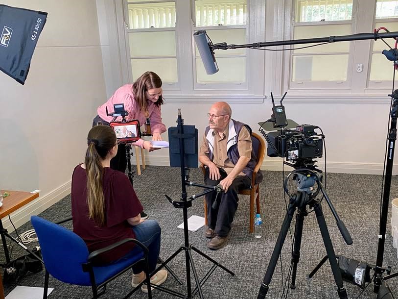 Image of three people - one filming a second person and thr=ird person leaning over and talking to second person